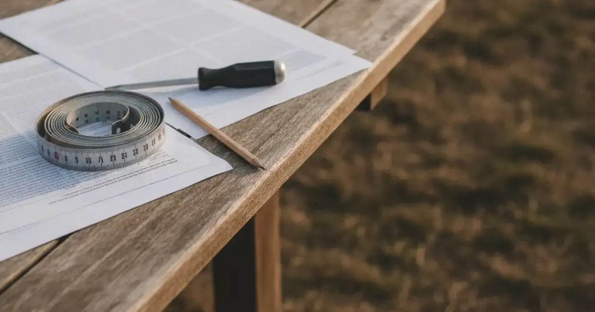 Devis et mètre ruban posés sur une table de jardin, terrain en préparation à l'arrière-plan flou